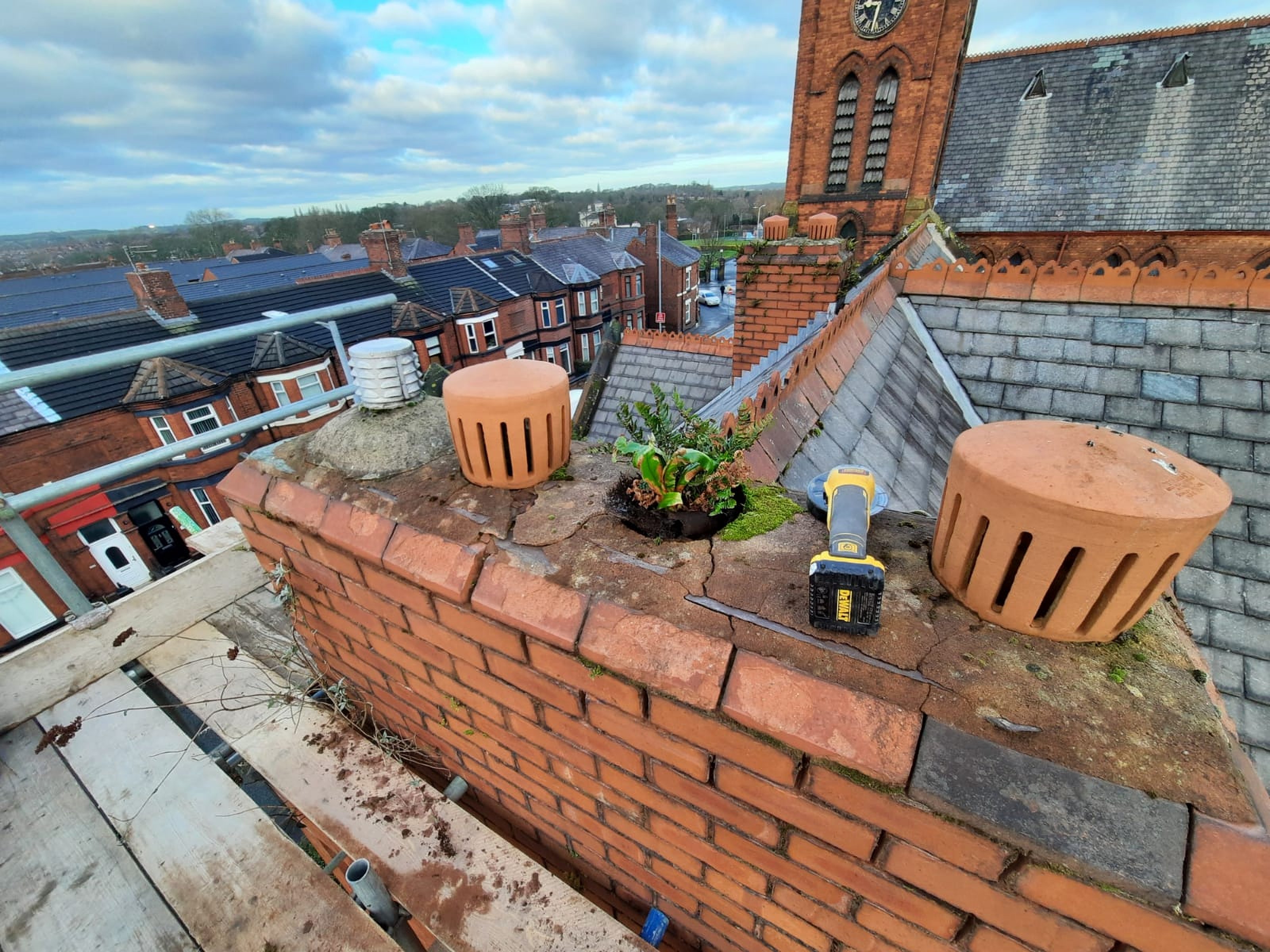 A high-angle shot of a brick chimney undergoing repair featuring terracotta chimney pots and scaffolding with a townscape in the background